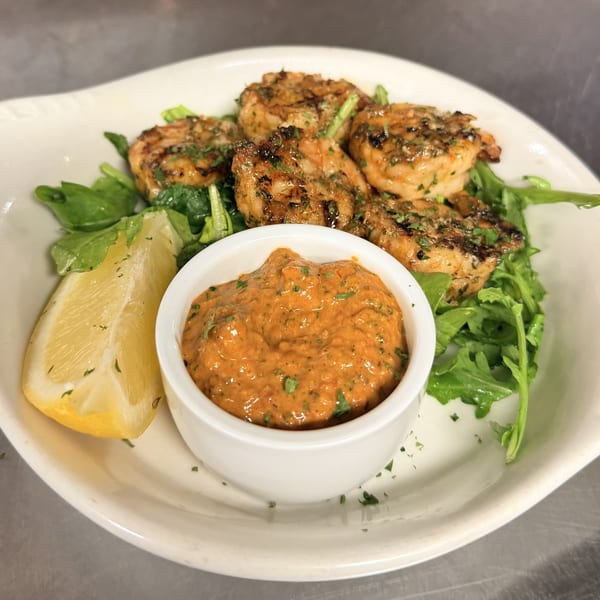 Grilled shrimp with fresh arugula, a lemon wedge, and a side of red dipping sauce on a white plate.
