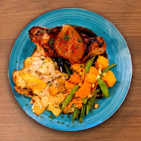 Grilled chicken with apricot glaze, scalloped potatoes, and mixed vegetables on a blue plate.