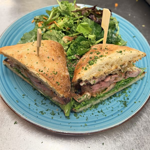 Steak sandwich cut in half with lettuce and onions, served with a green side salad on a blue plate.