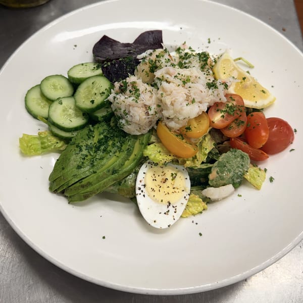 Colorful salad with crab, avocado, cucumber, tomatoes, egg, beets, and lemon wedge on a white plate.