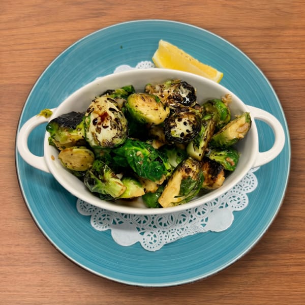 Roasted Brussels sprouts in a white bowl with a lemon wedge, on a blue plate with a doily.