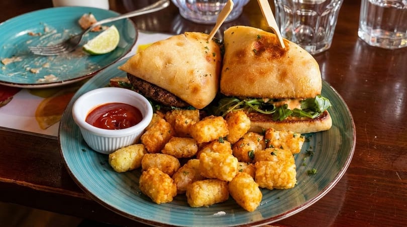 Two burgers with tater tots and ketchup on a blue plate, with an empty plate nearby.