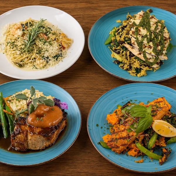 Four diverse gourmet dishes, including pasta, grilled chicken, pork chop, and salmon, arranged on a wooden table.