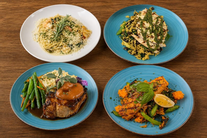 Four diverse gourmet dishes, including pasta, grilled chicken, pork chop, and salmon, arranged on a wooden table.