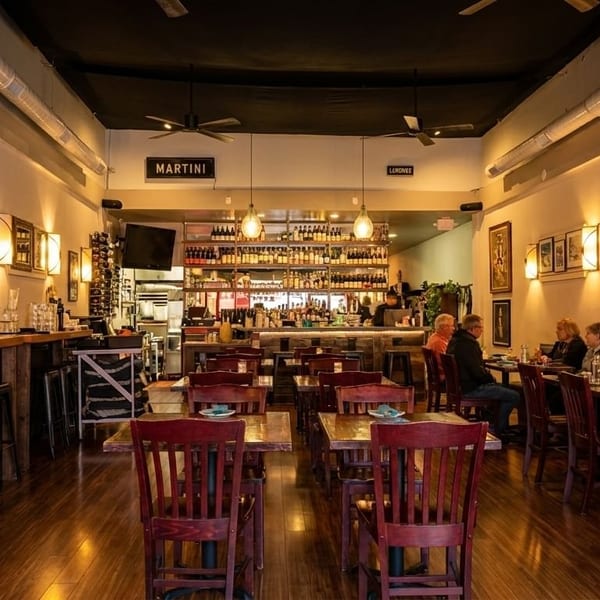 Restaurant interior with patrons dining, a long bar, and a wall with framed art and sconces.